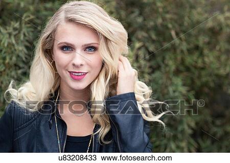 Stock Image - "Portrait of young blonde haired woman wearing leather jacket, hand in hair looking at camera smiling". Fotosearch