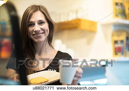 Stock Image - Portrait of young female waitress holding coffee and cake in cafe. Fotosearch