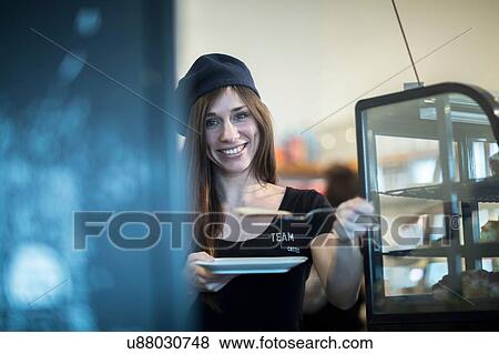 Stock Photo - Portrait of young female waitress holding plate and cake slice in cafe. Fotosearch