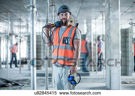 Stock Photography - Portrait of young male builder carrying circular saw on shoulder on construction site. Fotosearch