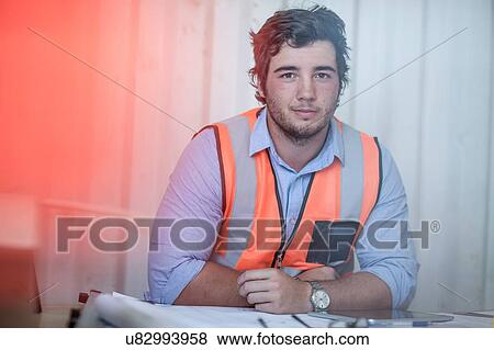 Stock Photo - Portrait of young male construction worker sitting at desk in portable cabin. Fotosearch