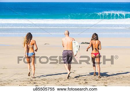 Stock Photography - Rear view of male surfer and two young women wearing swimwear on beach, El Cotillo, Fuerteventura, Spain. Fotosearch