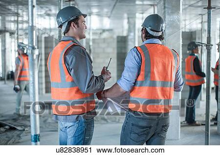 Stock Image - Rear view of site manager and builder meeting with blueprint on construction site. Fotosearch