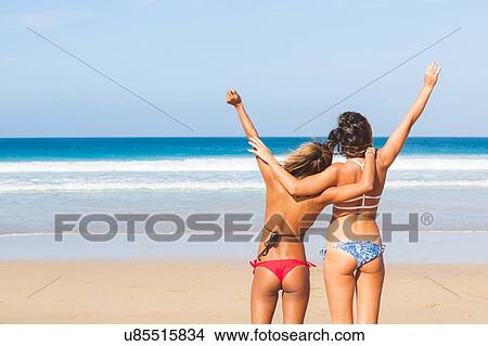 Picture - Rear view of two young women wearing bikinis with hands raised on beach, El Cotillo, Fuerteventura, Spain. Fotosearch
