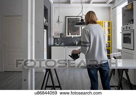 Stock Photo - Rear view of woman pouring tea from teapot at kitchen table. Fotosearch