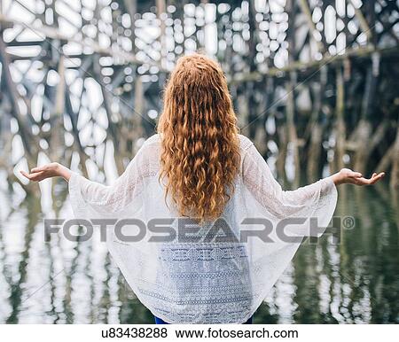Stock Photo - Rear view of woman with long red hair meditating in front of wood river pier. Fotosearch