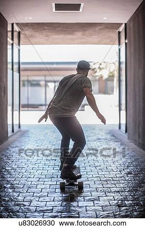 Stock Image - Rear view of young male skateboarder skateboarding in city. Fotosearch
