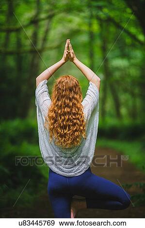 Stock Photo - Rear view of young woman in forest practicing yoga in tree pose. Fotosearch