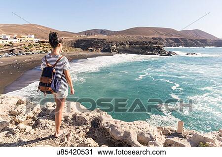 Stock Image - Rear view of young woman looking out at sea, Ajuy, Fuerteventura, Spain. Fotosearch