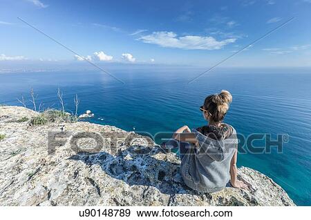 Rear view of young woman sitting on cliff looking out at view of ocean, Cagliari, Sardinia, Italy View Large Photo Image Stock Photo - Rear view of young woman sitting on cliff looking out at view of ocean, Cagliari, Sardinia, Italy. Fotosearch