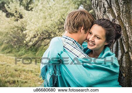 Stock Image - Romantic young couple leaning against tree wrapped in blanket. Fotosearch