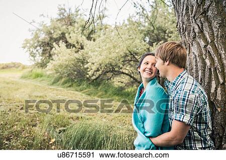 Stock Image - Romantic young couple leaning against tree. Fotosearch