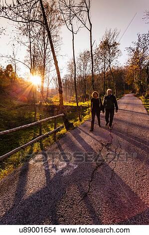 Picture - Senior couple strolling along sunlit rural road in aurumn, Lombardy, Italy. Fotosearch