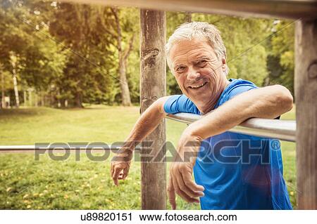 Stock Image - Senior man in park leaning against metal bar looking at camera smiling. Fotosearch