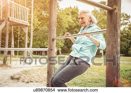 Picture - Senior woman in park doing limbo under metal bar smiling. Fotosearch