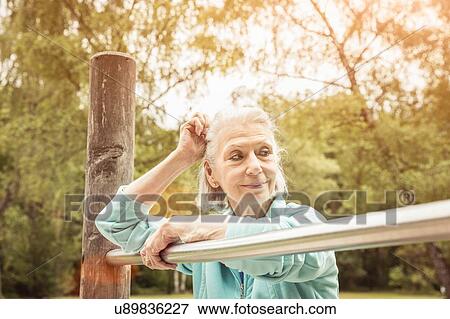 Stock Photo - Senior woman in park leaning against metal bar looking away smiling. Fotosearch