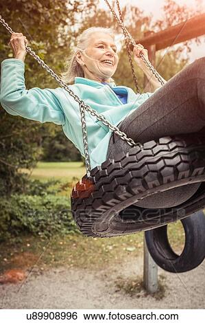 Stock Photograph - Senior woman swinging on tyre swing looking away smiling. Fotosearch
