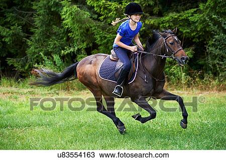 Stock Image - Side view of girl wearing riding hat galloping on horseback. Fotosearch