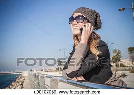 Stock Photo - Side view of young woman leaning against railings using smartphone smiling. Fotosearch