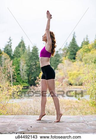 Stock Image - Side view of young woman practicing yoga on forest lakeside. Fotosearch