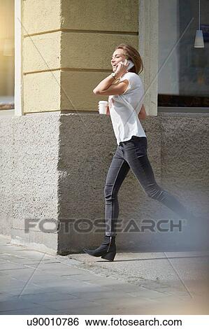 Stock Photograph - Side view of young woman running in street using smartphone to make telephone call. Fotosearch
