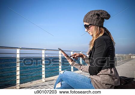 Side view of young woman sitting by seafront using digital tablet View Large Photo Image Stock Image - Side view of young woman sitting by seafront using digital tablet. Fotosearch