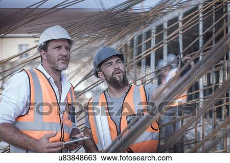 Stock Image - Site manager and builder discussing steel rod structures on construction site. Fotosearch