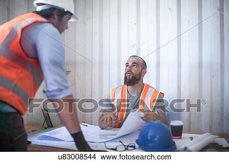 Picture - Site manager sitting at desk discussing blueprint with worker in portable cabin. Fotosearch