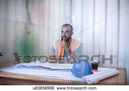 Stock Photo - Site manager sitting at desk with blueprint in portable cabin. Fotosearch