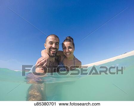 Stock Image - Surface level view of young couple taking selfie in sea. Fotosearch