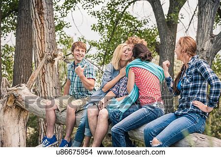 Picture - Teenage girl and young woman kissing young man on cheek whilst sitting on fallen tree. Fotosearch