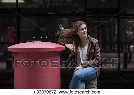 Stock Image - Teenage girl with flyaway hair sitting next to red city post box. Fotosearch