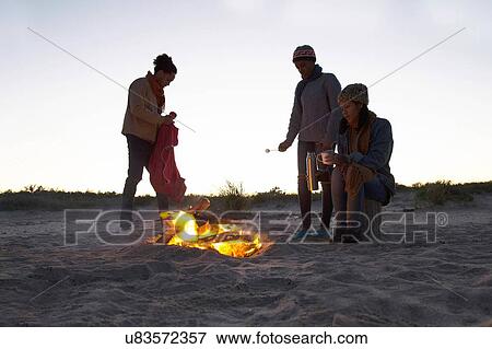 Stock Photo - "Three friends on beach, gathered around camp fire". Fotosearch
