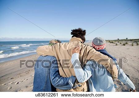 Stock Image - "Three friends walking along beach, arms around each other, rear view". Fotosearch