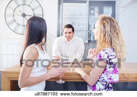 Stock Image - Three young adult friends chatting and drinking wine in kitchen. Fotosearch
