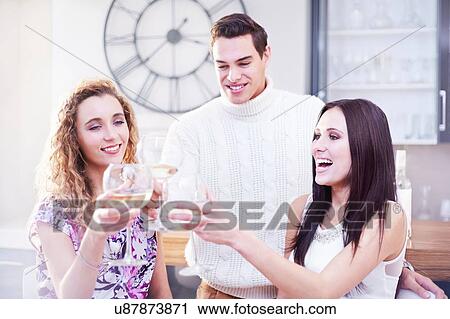 Stock Image - Three young adult friends making a white wine toast in kitchen. Fotosearch