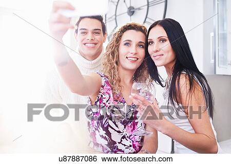Stock Photograph - Three young adult friends posing for smartphone selfie in kitchen. Fotosearch