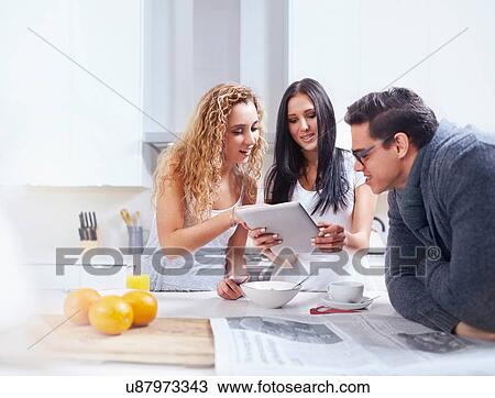 Stock Image - Three young adults using digital tablet at kitchen counter. Fotosearch
