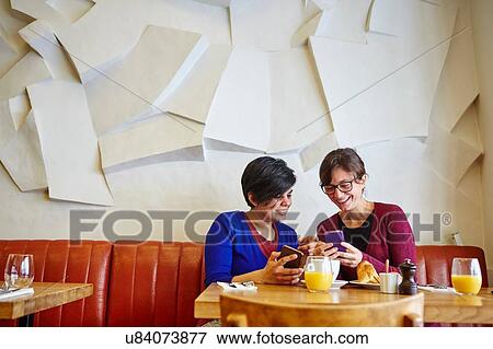 Stock Photo - Two adult female friends reading smartphone text in restaurant. Fotosearch