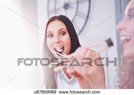 Stock Photo - Two female friends eating sushi with chopsticks in kitchen. Fotosearch