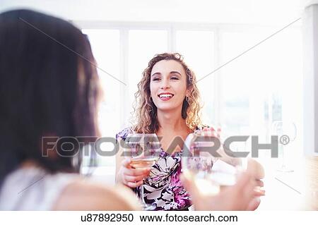 Stock Image - Two female friends making a white wine toast in kitchen. Fotosearch