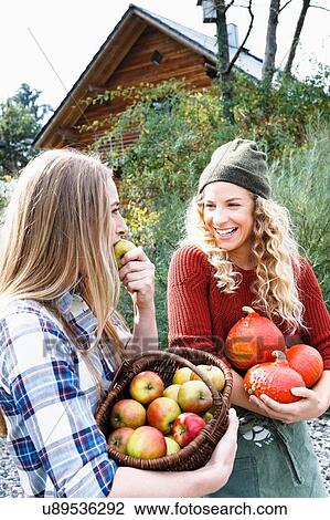 Stock Image - Two friends carrying homegrown produce, one woman eating apple. Fotosearch
