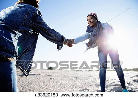 Stock Image - "Two friends fooling around at beach, holding hands, smiling". Fotosearch