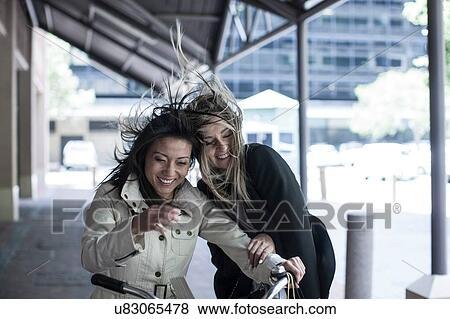 Two women friends with flyaway hair laughing on bicycle in city View Large Photo Image Stock Photo - Two women friends with flyaway hair laughing on bicycle in city. Fotosearch