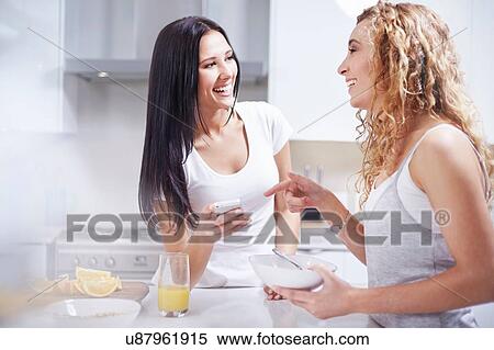 Stock Photography - Two young female friends laughing at kitchen counter. Fotosearch