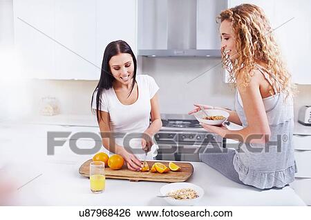 Stock Photograph - Two young female friends preparing breakfast oranges at kitchen counter. Fotosearch