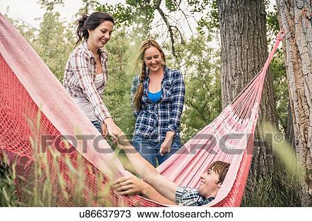 Stock Image - Two young women helping young man from hammock. Fotosearch
