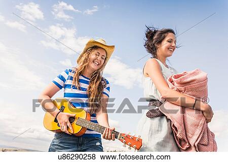 Stock Photo - Two young women playing ukulele and carrying blanket against blue sky. Fotosearch