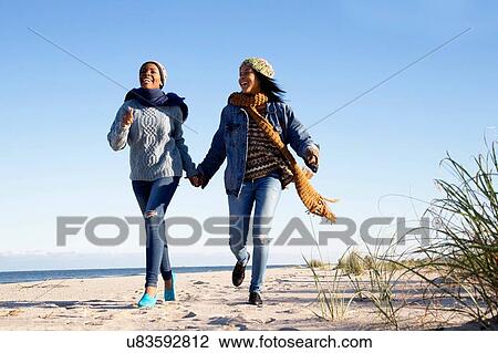 Stock Image - "Two young women, running along beach, holding hands, laughing". Fotosearch