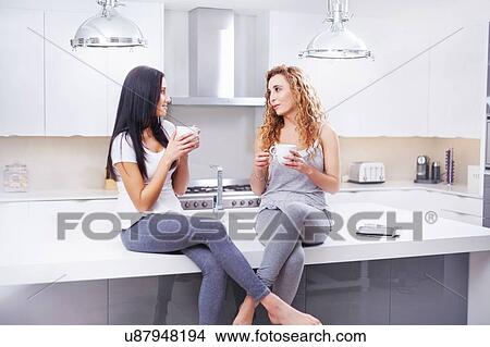 Two young women sitting on kitchen counter gossiping and drinking coffee View Large Photo Image Picture - Two young women sitting on kitchen counter gossiping and drinking coffee. Fotosearch